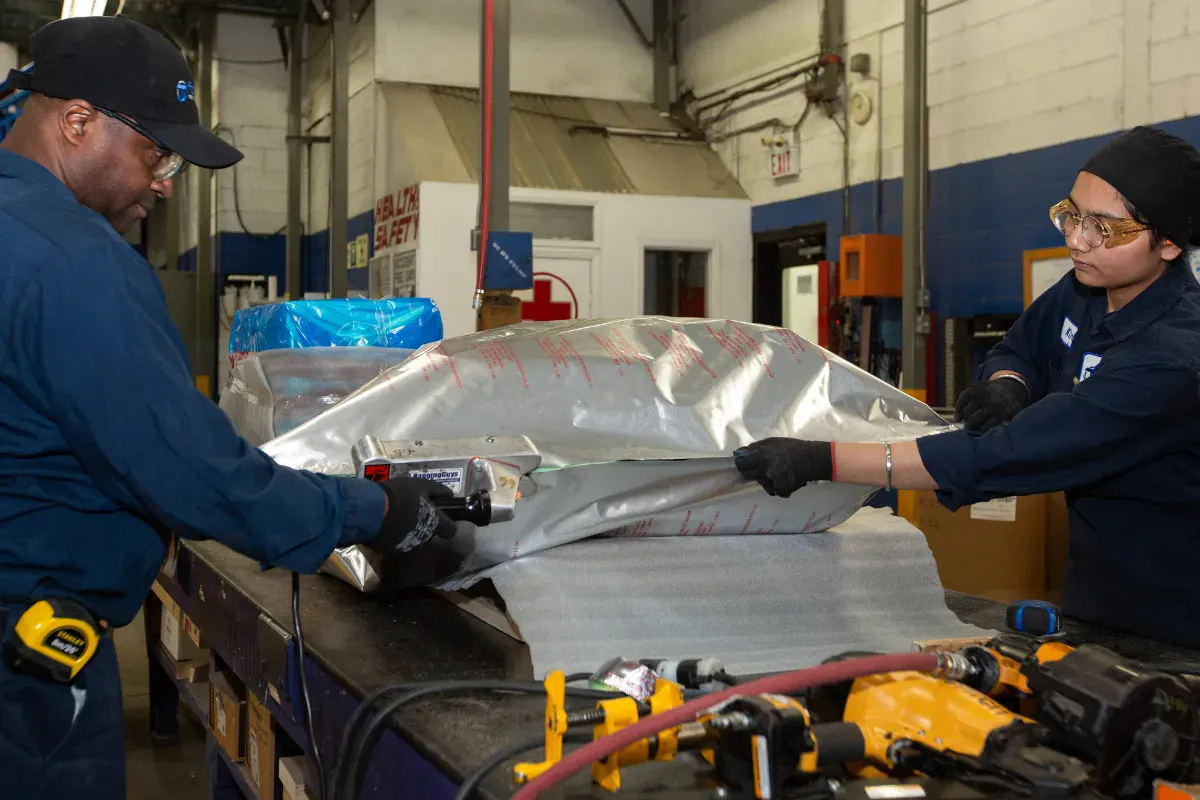 Two workers heat sealing barrier foil around equipment for corrosion protection packaging.