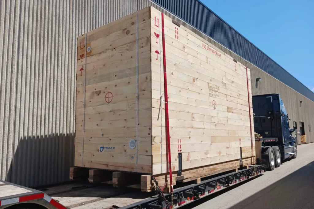 Oversized wooden export crate secured to a flatbed trailer outside an industrial facility
